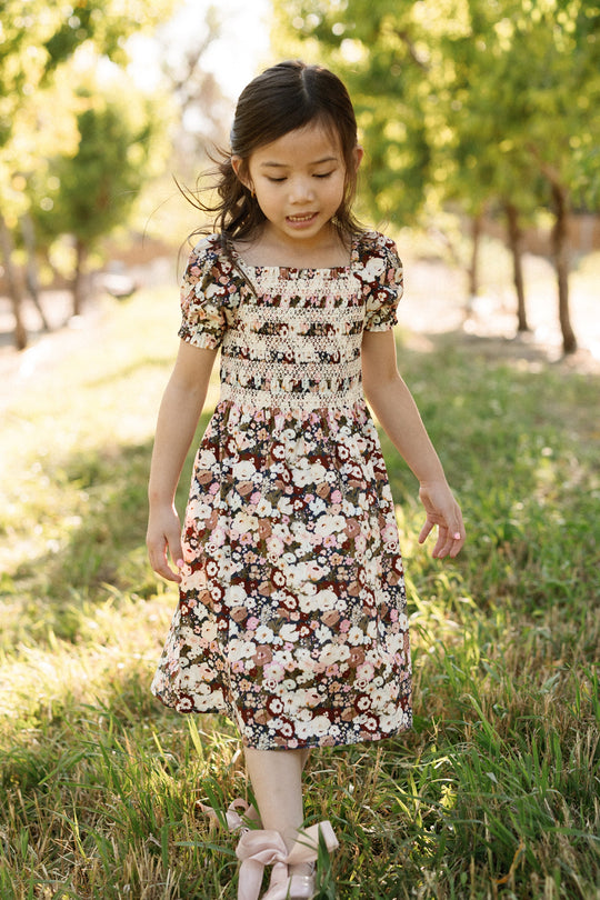 A young girl in the Ivy City Co Mini Sunday Best Dress in Midnight Garden, featuring puff sleeves and a floral pattern, walks outdoors on grass surrounded by greenery and sunlight. She looks down with a gentle smile, her loose hair complemented by large pink bows on her shoes.