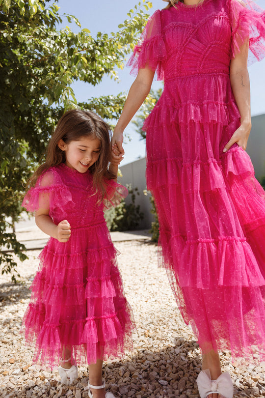 A young girl and an adult, both wearing matching Ivy City Co Mini Whimsical Dresses in Fuchsia featuring flutter sleeves and layered tulle skirts, hold hands and walk outside on a sunny day, surrounded by greenery and gravel.