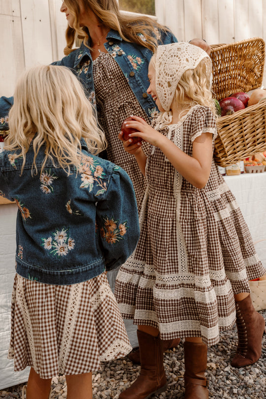 Three girls wearing the Ivy City Co Mini Palermo Dress in Hickory Gingham stand outdoors by a basket of apples. Two, with blonde hair, layer denim jackets with floral embroidery over their dresses; one holds an apple while another wears a lace headscarf and brown boots. The charming dresses add a vintage touch to their playful afternoon.
