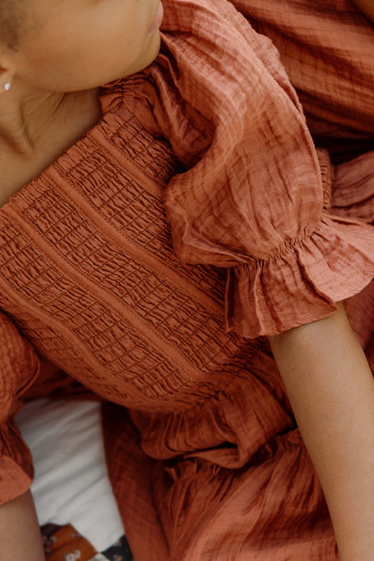 A child wearing the Ivy City Co Mini Madeline Dress in Apple Cider, featuring a textured fabric, smocked bodice, and ruffled sleeves, sits closely next to another person dressed similarly; both are partially visible.