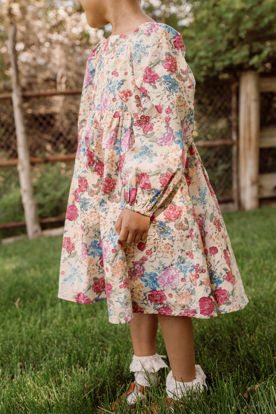 A young child wearing the Ivy City Co Mini Adelpha Dress in Autumn Amity Floral, featuring balloon sleeves and a smocked bodice, stands on green grass outdoors. The background includes a wooden fence and trees, with only the lower face and body visible.