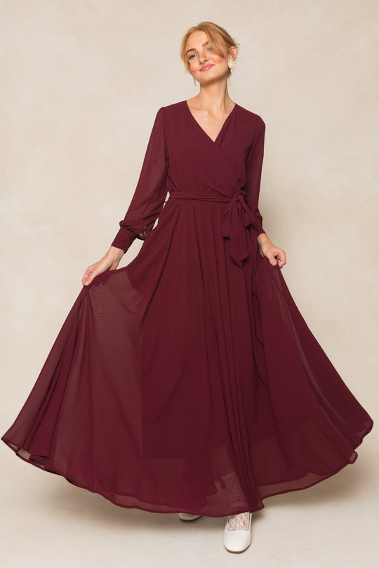 A woman stands against a light beige background, gently holding the sides of her Ivy City Co Andie Dress in Wine Chiffon—a long-sleeved maxi dress with a flowing skirt. She smiles softly, her hair styled up, and pairs the look with white shoes.