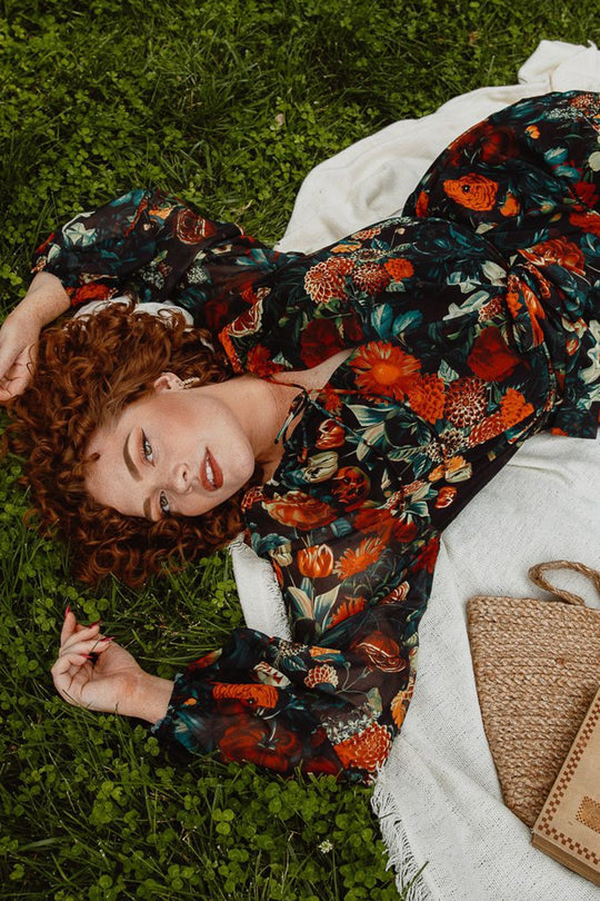 A woman with curly red hair lies on the grass atop a white blanket, wearing Ivy City Co’s Imogen Dress in Moody Floral—a long-sleeve, elegant, and maternity-friendly maxi dress. She gazes upward with a slight smile while a woven handbag rests nearby on the blanket.