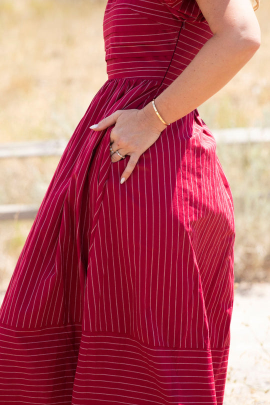 A person wearing the Ivy City Co Julie Dress in Cranberry Pinstripe, which features puff sleeves and a smocked bodice, stands outdoors with one hand on their hip. Only their torso and arm are visible against a blurred natural backdrop.