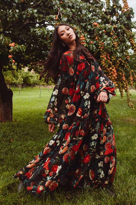 A woman in a flowing Imogen Dress in Moody Floral by Ivy City Co stands gracefully on grass beneath a fruit-laden tree, surrounded by greenery.