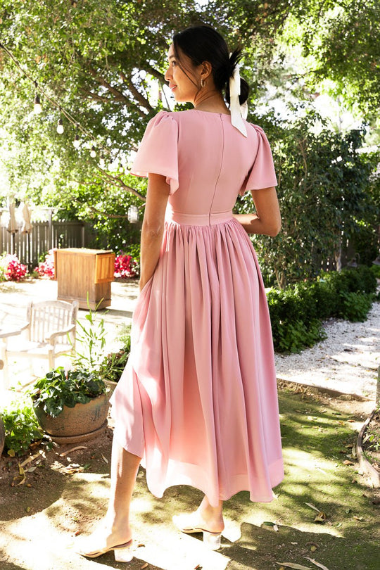 A woman wearing the Abbie Dress in Blush by Ivy City Co, featuring flutter sleeves, stands outdoors in a sun-dappled garden with her back turned. A white ribbon adorns her hair and she pairs the dress with light-colored flats. Elegant trees and garden furniture complete the scene, highlighting this modest bridesmaid dress.