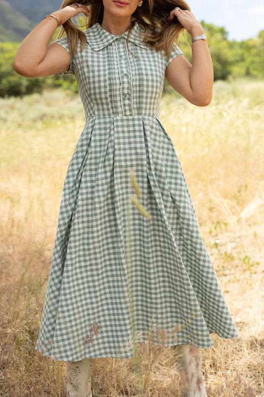 A woman wearing the Amber Dress in Sage Gingham by Ivy City Co stands in a grassy field, holding her hair. The cotton midi dress features short sleeves, a collar, and button-down front detailing. Trees and hills appear in the background.