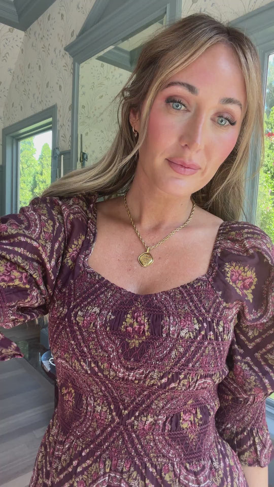 A woman with long blonde hair and light eyes is wearing the Josephine Dress in Nocturne Diamond Rose by Ivy City Co—a patterned maroon cotton dress featuring a smocked bodice and paired with a gold necklace. She stands indoors near windows overlooking lush greenery, gazing serenely at the camera. This maternity-friendly outfit blends comfort with elegance.