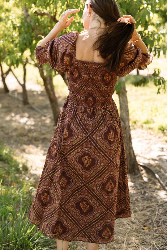 A woman stands outdoors in the Josephine Dress in Nocturne Diamond Rose by Ivy City Co, the maroon patterned cotton fabric and smocked bodice visible as she faces away and adjusts her hair amidst trees and warm sunlight.