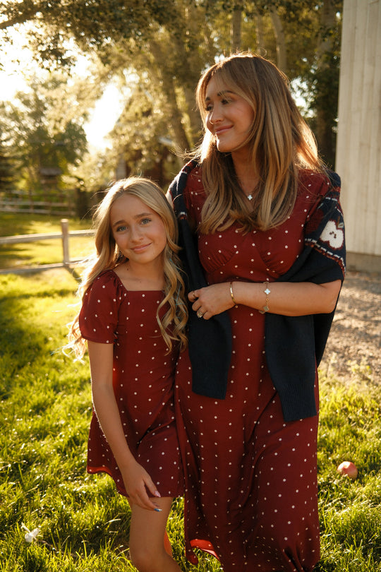 Two people wearing matching Ivy City Co Teen Blakely Dresses in Cranberry Dainty Dot with petal sleeves stand close together outside, smiling in the sunlight, with green grass and trees in the background.