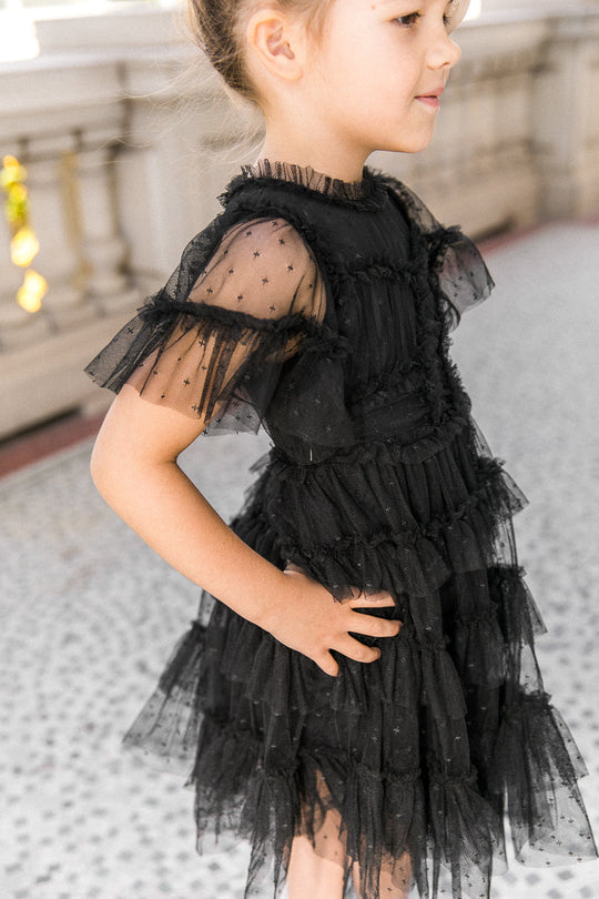 A young girl stands sideways with hands on hips near a stone railing, wearing the Ivy City Co Mini Whimsical Dress in Black, which features a layered tulle skirt and sheer, star-patterned sleeves.