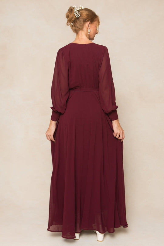 A woman with light hair styled in an updo adorned with small flowers wears the Ivy City Co Andie Dress in Wine Chiffon, a long-sleeved, floor-length gown. She stands with her back to the camera against a neutral background, highlighting this elegant bridesmaid-ready look.