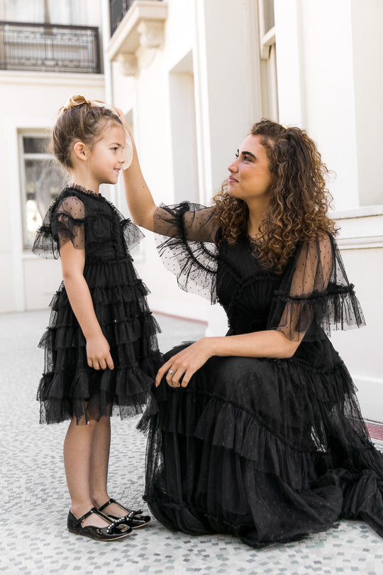A woman and a young girl gently touch heads as they stand outside a light-colored building, both wearing the Ivy City Co Mini Whimsical Dress in Black. Their matching dresses feature ruffled skirts, flutter sleeves, and delicate layered tulle.