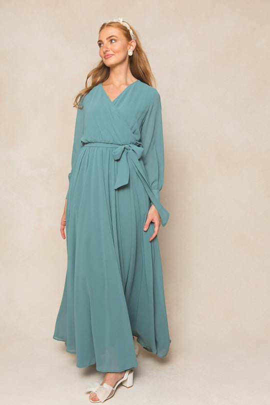 A woman wears the Andie Dress in Seaglass Chiffon by Ivy City Co—a long-sleeve wrap dress featuring a waist tie—as she stands against a neutral background, accessorized with white sandals and a headband, gazing slightly to the side with a gentle smile.