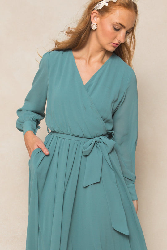 A woman with long hair, accessorized with earrings and a white headband, looks down with one hand in her pocket while wearing the Ivy City Co Andie Dress in Seaglass Chiffon— a teal, long-sleeved, maxi-length wrap dress featuring a waist tie and pockets.
