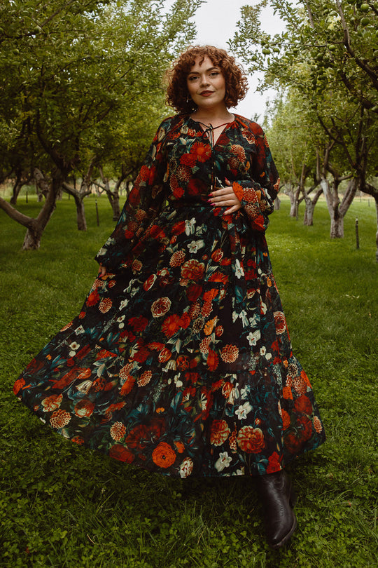 Clad in the Imogen Dress in Moody Floral by Ivy City Co—a stylish, maternity-friendly floral maxi—a curly-haired person pairs it with black boots while standing on lush green grass amid leafy orchard trees.