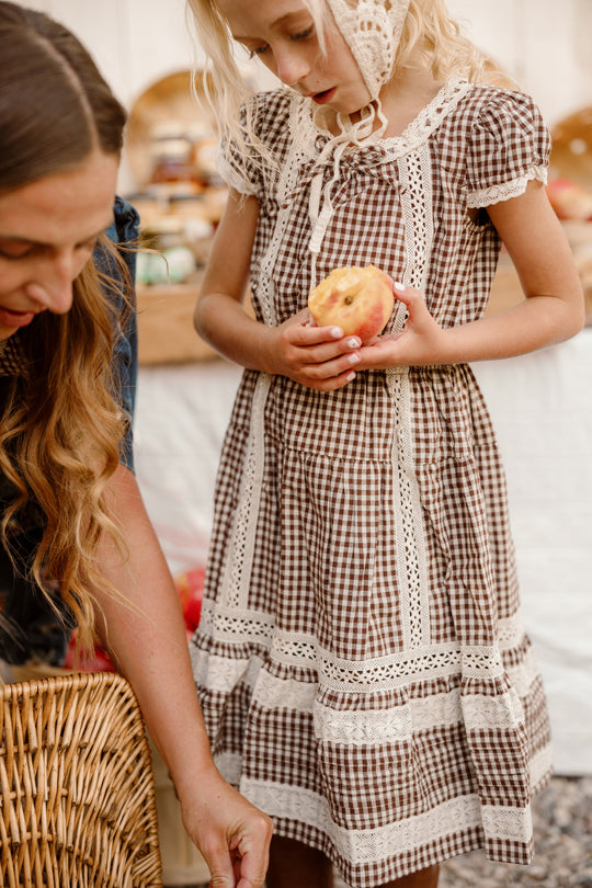 A young girl wearing the Ivy City Co Mini Palermo Dress in Hickory Gingham, featuring lace trim, holds a partially eaten apple while standing next to a woman with long wavy hair at a market or picnic setting.