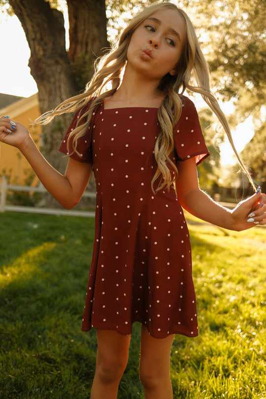 A young girl with long blonde hair stands outdoors on grass, playfully holding her hair and making a duck face as sunlight filters through the trees. She is wearing the Ivy City Co Teen Blakely Dress in Cranberry Dainty Dot, a charming midi-length piece featuring petal sleeves and a delightful red polka dot pattern.