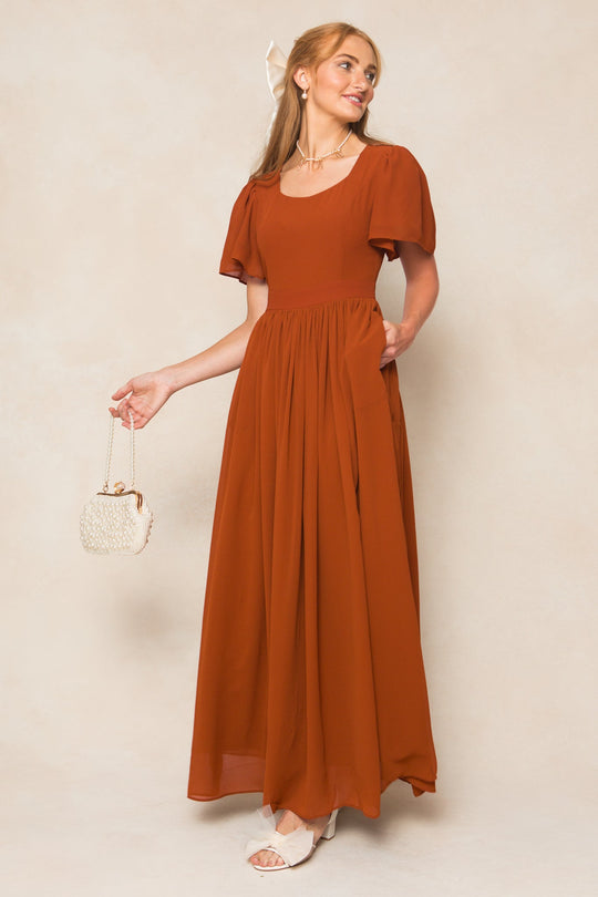 A woman wearing the Ivy City Co Abbie Dress in Amber Chiffon—a flowing, maxi-length dress with short puff sleeves—holds a small woven handbag, pairs it with white sandals, and smiles while looking to the side against a light background.