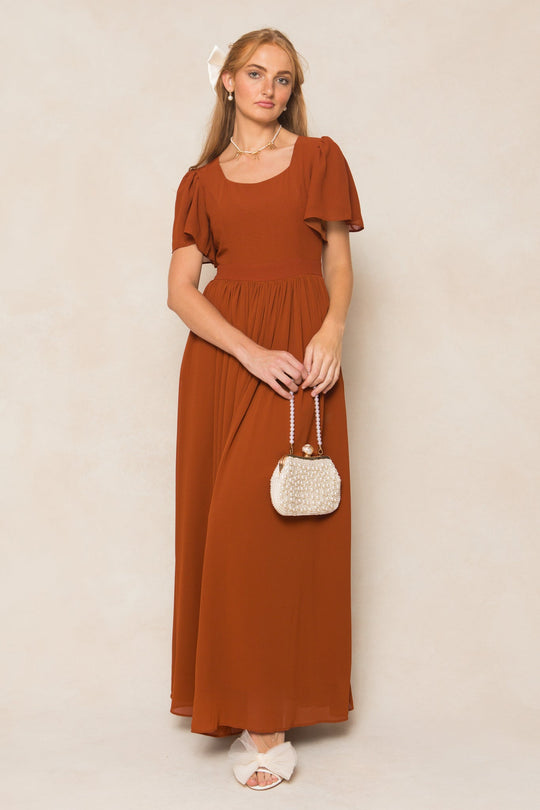 A woman wears the Abbie Dress in Amber Chiffon by Ivy City Co—a rust-colored, short-sleeved maxi dress—pairing it with a small pearl-beaded purse, white sandals, and a large white bow in her hair as she stands against a light beige background.