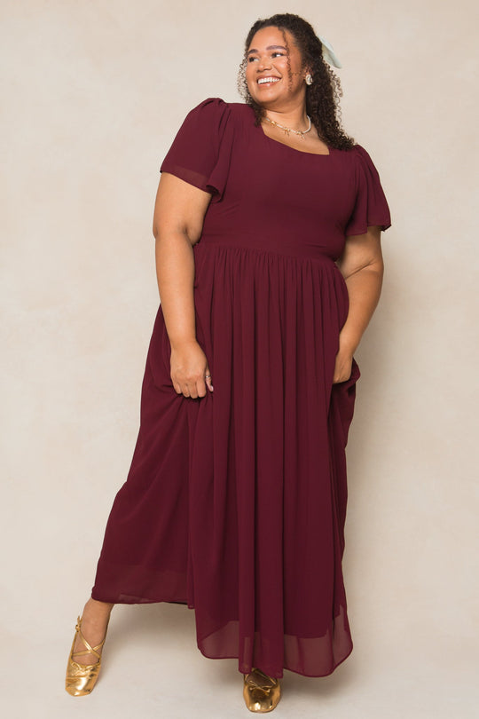 A smiling woman wears the Ivy City Co Abbie Dress in Wine Chiffon, a short-sleeve, floor-length bridesmaid gown, paired with gold shoes. She stands against a beige background, looking to the side with one hand in her pocket and the other by her side.