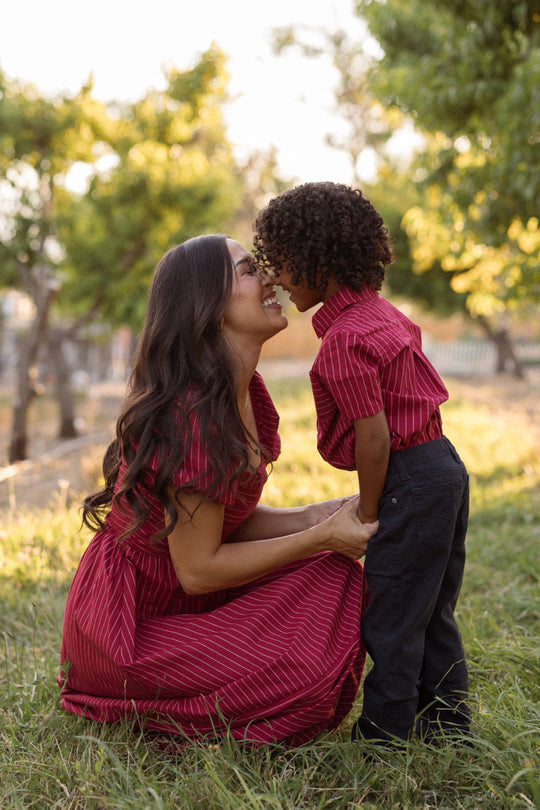 A woman kneels in the grass, smiling as she touches noses with a young boy. Both are dressed in matching Ivy City Co Julie Dresses in Cranberry Pinstripe, featuring a smocked bodice and charming puff sleeves. Trees and sunlight fill the background.