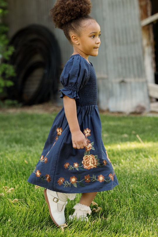 A young girl with curly hair, dressed in the Ivy City Co Mini Teagan Dress in Navy Embroidered Fall Floral—which features a smocked bodice and delicate floral embroidery—and white shoes, walks on green grass beside a corrugated metal building.
