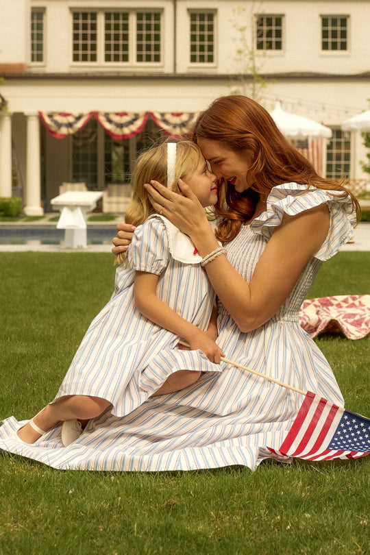 A woman and young girl wear matching Ivy City Co Hattie Dresses in Heritage Stripe as they kneel on the grass, smiling with foreheads touching. The girl holds an American flag near a house adorned with patriotic bunting in the background.