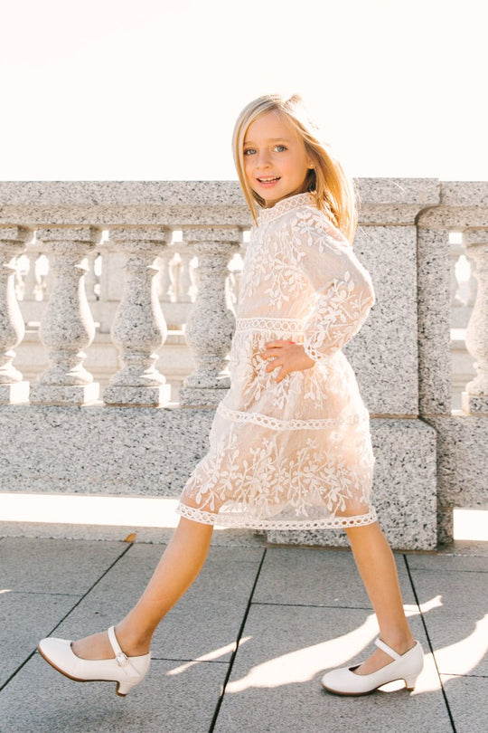 A smiling young girl walks outdoors on a sunny day, wearing the Ivy City Co Mini Sicily Dress with Long Sleeves and white shoes, with a stone railing in the background.