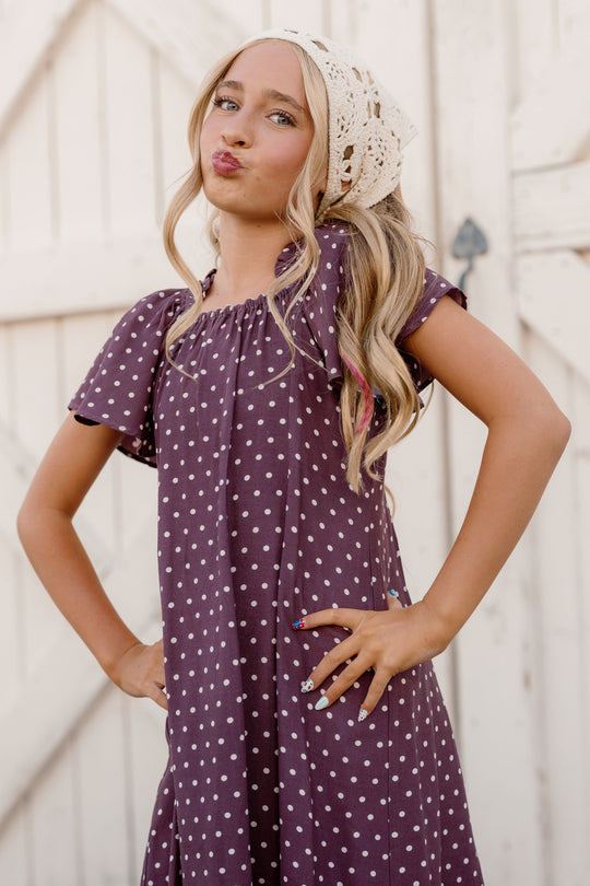 A girl poses confidently in the Ivy City Co Teen Blakely Dress in Aubergine Ditsy Dot, featuring flutter sleeves and paired with a crocheted headscarf, standing in front of a white wooden fence with her hands on her hips and lips puckered.