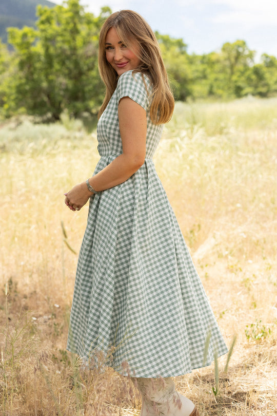 A woman wearing the Ivy City Co Amber Dress in Sage Gingham, featuring a pleated skirt, stands in a sunlit field with trees and grass in the background, looking over her shoulder and smiling.
