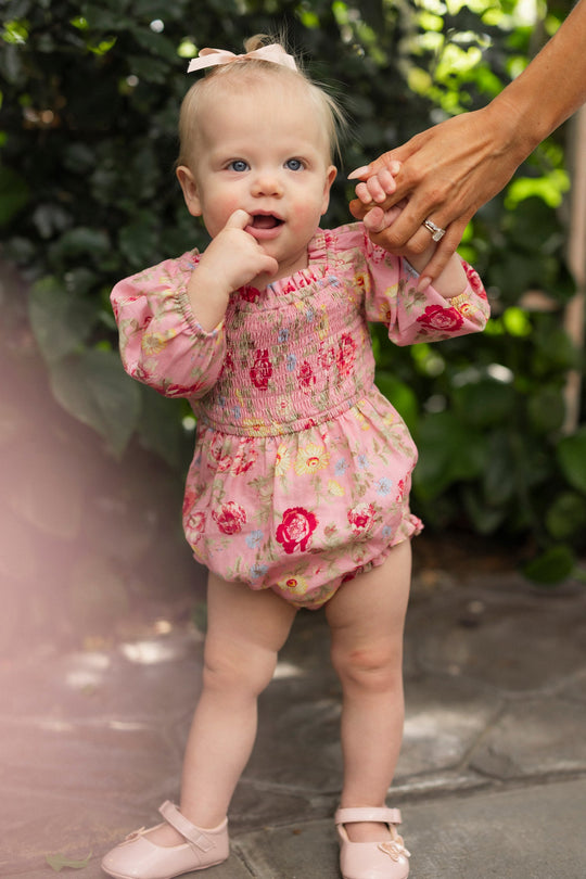 A baby with light hair, wearing a bow and matching pink shoes, stands on a stone path surrounded by lush greenery. She is dressed in the Ivy City Co Baby Madeline Romper in Pink Vintage Poppies, featuring a smocked bodice and charming floral print, while holding an adult’s hand and touching her mouth.