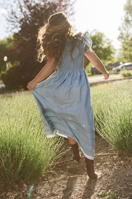 A young girl with long, wavy hair wears the Ivy City Co Hattie Ankle Length Dress in Embroidered Chambray as she runs through a sunlit field, holding up her skirt while following a dirt path surrounded by tall grass.