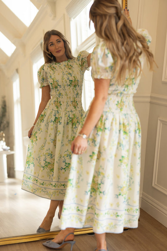 Wearing the Jennifer Dress in Garden Tea Party Floral by Ivy City Co, a woman with puff sleeves admires her reflection in a tall mirror in a bright, elegant room with wooden floors and white walls.