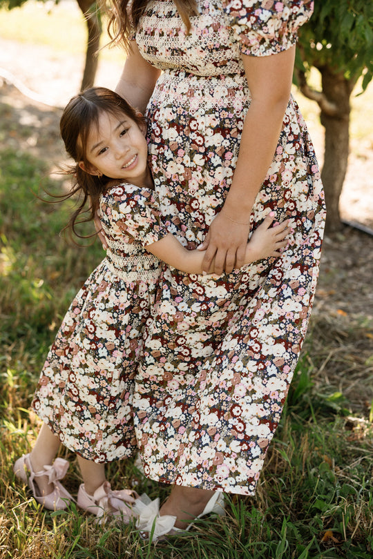 A young girl hugs a woman’s waist outdoors, both standing on grass near trees in their matching Ivy City Co Mini Sunday Best Dress in Midnight Garden—styled with smocked bodices and charming puff sleeves.