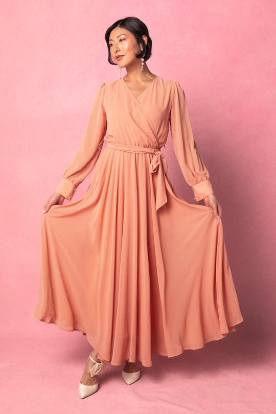 A woman stands against a pink background in the Ivy City Co Andie Dress in Apricot Crush, featuring long sleeves and a wrap silhouette. She holds out the sides of her flowing maxi skirt, accessorized with cream heels and dangling earrings—an elegant option for both everyday wear and maternity style.