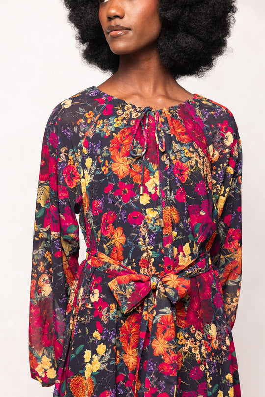 A woman with curly hair, her face partially visible, wears the Imogen Dress in Night Garden by Ivy City Co—a long-sleeve maxi dress with a striking fall floral print featuring red, yellow, and orange flowers. Designed to be maternity friendly, the style features elegant details like a bow at the waist and a keyhole neckline, blending comfort and sophistication for any occasion.