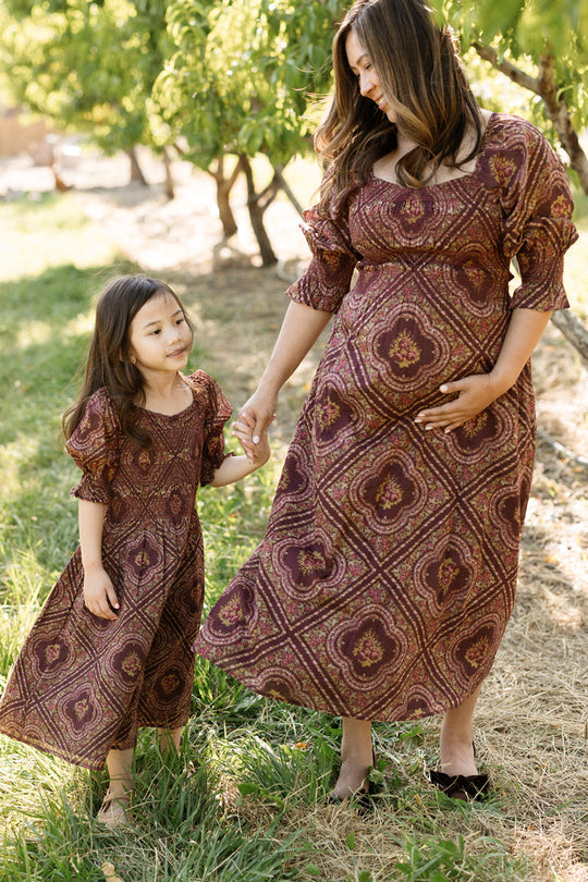 A woman and a young girl hold hands outdoors, both wearing the Ivy City Co Josephine Dress in Nocturne Diamond Rose. Featuring smocked bodices, these maternity-friendly cotton dresses keep them comfortable as they smile together on the grass near trees, enjoying a sunny day.