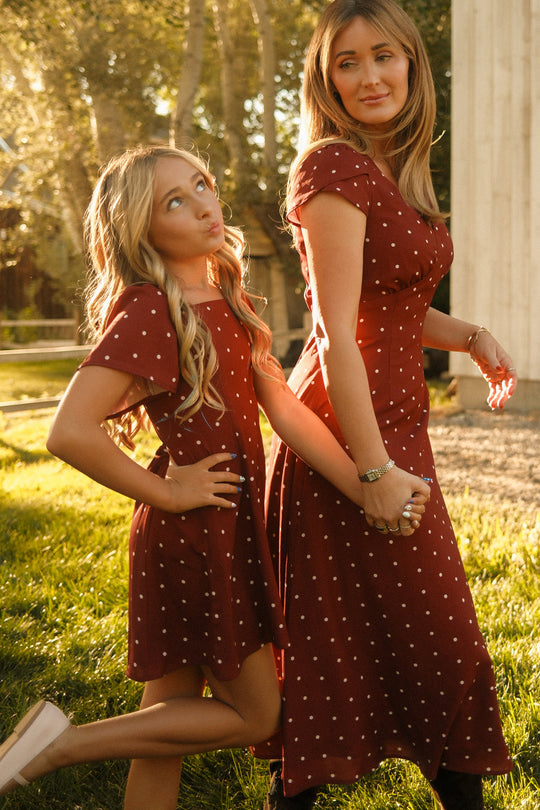 A woman and a girl stand outdoors, holding hands and both wearing coordinated maroon polka dot dresses. The girl poses playfully in the adorable Ivy City Co Teen Blakely Dress in Cranberry Dainty Dot, featuring charming petal sleeves, while the woman glances back with a warm smile. Sunlight filters through the trees behind them, accentuating the elegant midi-length silhouettes of their outfits.