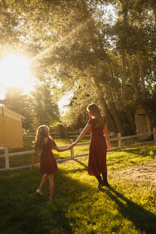 Two girls in matching Ivy City Co Teen Blakely Dresses in Cranberry Dainty Dot, featuring delicate petal sleeves, hold hands as they walk on grass under golden sunlight, with trees and wooden fences surrounding them and creating a warm, serene outdoor scene.