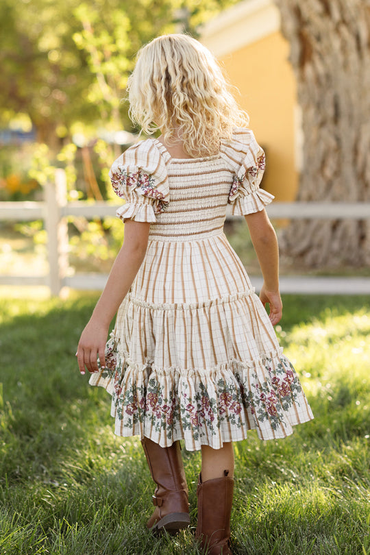 A young girl with blonde curly hair wears the Ivy City Co Mini Madeline Dress in Embroidered Cider Grid, featuring puff sleeves and a smocked bodice, as she strolls across green grass near a wooden fence and a large tree, sunlight filtering behind her. She completes her charming look with brown boots.