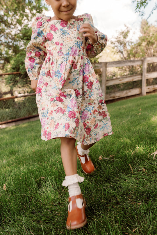 A child wearing the Mini Adelpha Dress in Autumn Amity Floral by Ivy City Co, with its smocked bodice and charming balloon sleeves, smiles while walking on green grass, with a wooden fence and trees in the background.