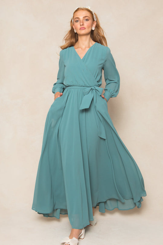 A woman poses against a beige background wearing the Andie Dress in Seaglass Chiffon by Ivy City Co—a flowing, light teal maxi-length chiffon dress featuring long sleeves, a tied waist, and convenient pockets. She completes the look with white heels, pearl hair accessories, and her long hair styled down. This elegant dress is an ideal choice for fall bridesmaids or any special occasion.