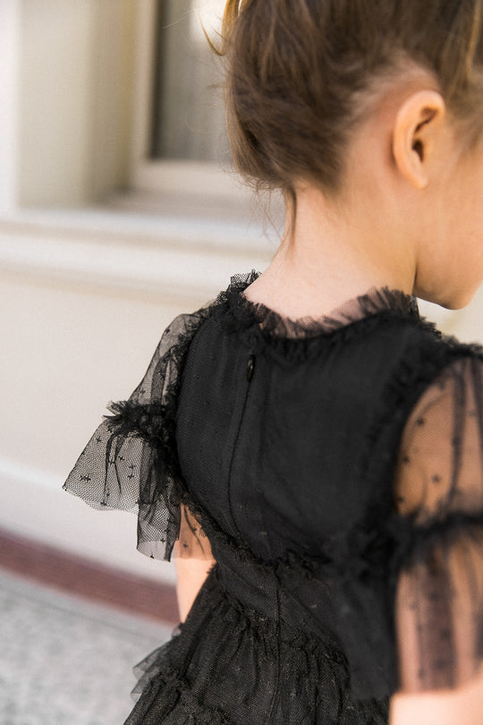 A young child facing away wears the Ivy City Co Mini Whimsical Dress in Black, featuring ruffled short sleeves and sheer polka-dot fabric on the sleeves and back. The layered tulle skirt adds playful volume, set against a backdrop of a cream-colored wall and part of a window.