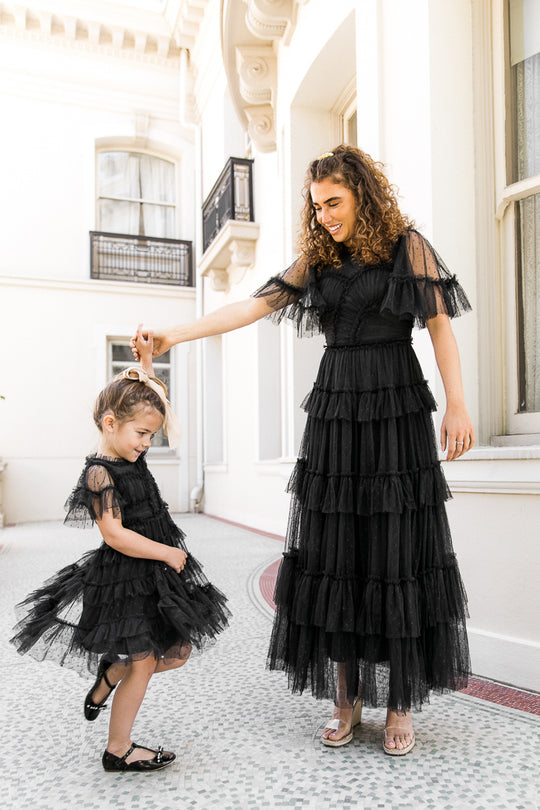 A woman and a young girl, both dressed in matching Ivy City Co Mini Whimsical Dresses in Black featuring ruffled details and layered tulle skirts, stand outside a building. The woman twirls the girl, who smiles and looks down, both radiating joy and elegance.