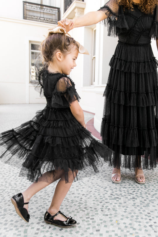 A young girl twirls in the Ivy City Co Mini Whimsical Dress in Black, its ruffled bodice and layered tulle skirt matching an adult companion’s dress. They hold hands in a bright room with white walls and a tiled floor.