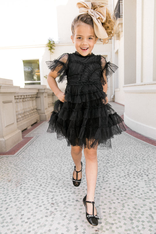 A young girl walks and smiles outdoors on a tiled pathway near a white building, wearing the Ivy City Co Mini Whimsical Dress in Black with a layered tulle skirt, paired with black shoes and a large beige bow in her hair.