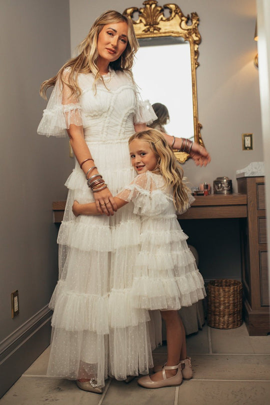 A woman and a young girl wear matching Ivy City Co Whimsical Dresses in White, standing together in a warmly lit room. The girl hugs the woman as a gold ornate mirror hangs on the wall behind them.