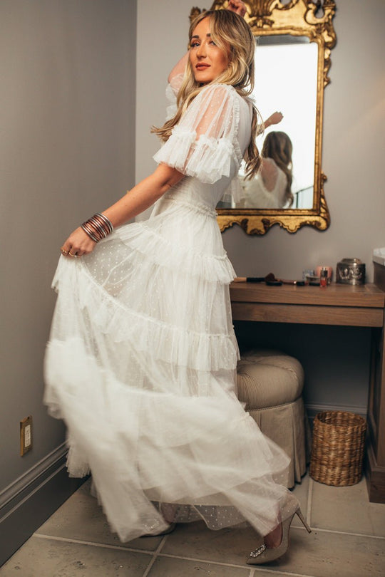A woman models the Ivy City Co Whimsical Dress in White indoors, lifting the elegant skirt. She pairs it with metallic heels and bracelets. Her long wavy hair complements the scene, with a mirror, vanity, and stool in the background.