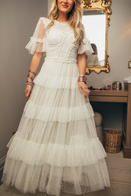 A woman stands in a softly lit room before a mirror and wooden vanity, wearing the Ivy City Co Whimsical Dress in White—featuring a ruffled tulle skirt and sheer short sleeves. Her long, wavy blonde hair falls over bangles on her wrist.